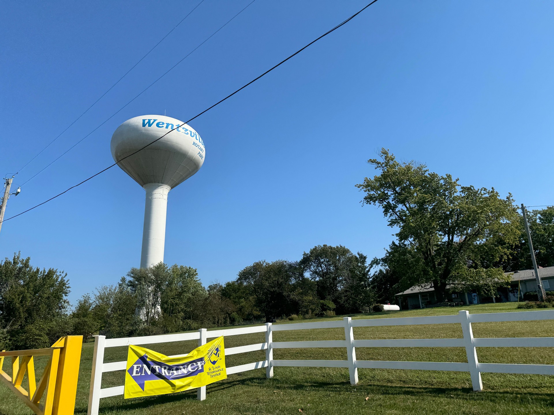 Wentzville water tower stands prominently against clear blue skies near a white fence